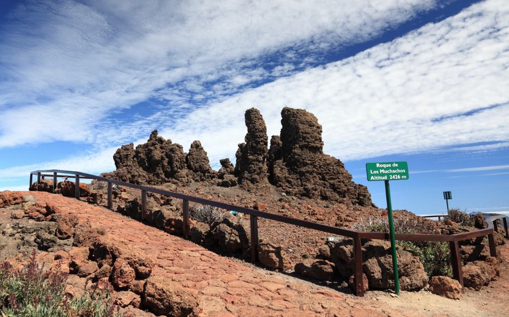 At the summit of Roque de Los Muchachos in La Palma, Canary Islands, rugged volcanic rock formations stand against a bright blue sky, with an informative sign indicating the location and altitude of 2426 meters.
