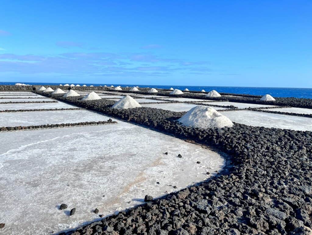 A vibrant view of Salinas de Fuencaliente La Palma, showcasing a patchwork of salt pans bordered by black volcanic rocks, with mounds of harvested salt under the bright blue sky.