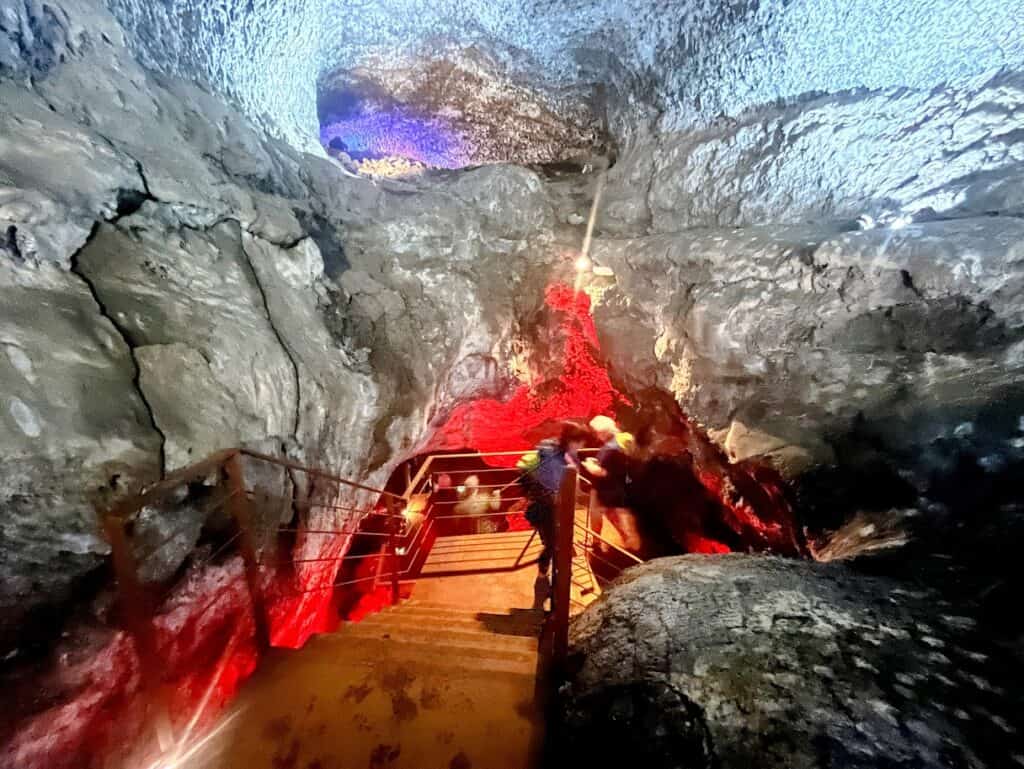Visitors navigate down a well-maintained staircase within the volcanic cave, surrounded by the dramatic textures of the ancient lava flow at Caños de Fuego.