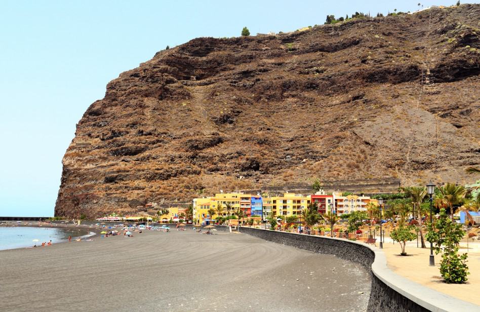 The serene black sands of Tazacorte Beach when you visit La Palma, Canary Islands, lie beneath dramatic cliffs, with colorful buildings in the background, beckoning beachgoers and sun worshippers looking for tranquil things to do in La Palma.