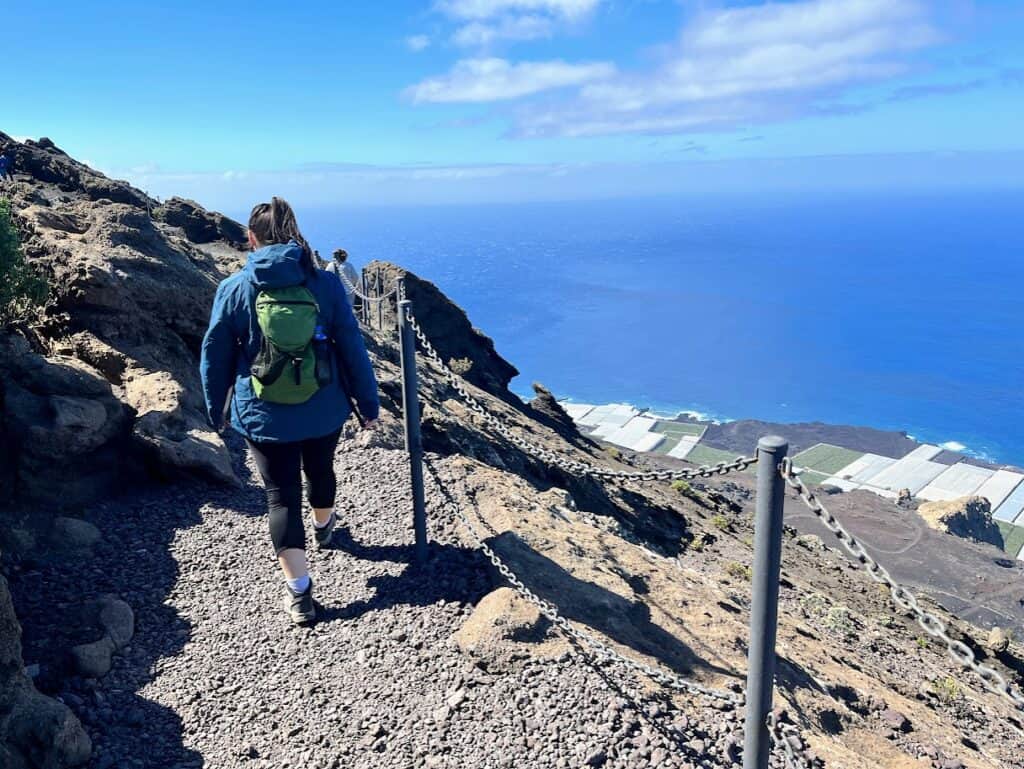 A hiker traversing the rugged trail on a Volcán San Antonio hike, with the ocean and cultivated land in the distance, showcasing the adventurous activity available to visitors in La Palma.