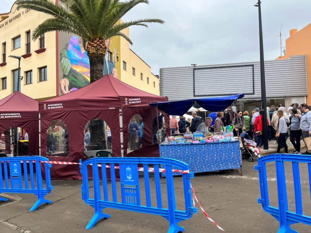 A lively street festival in La Palma with people browsing stalls under maroon tents, showcasing local crafts and products, with a mural and palm tree in the background.