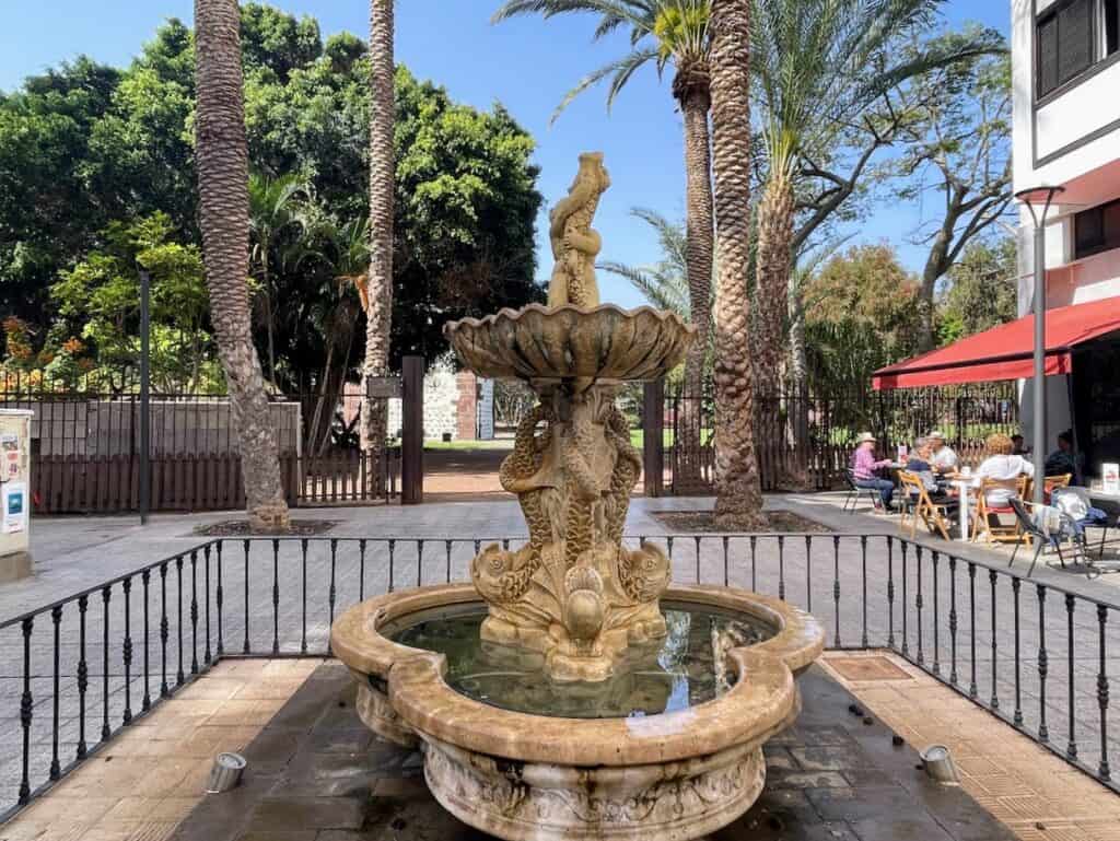 A charming stone fountain in San Sebastián de La Gomera, with palm trees and a café terrace in the background – a serene spot to add to your La Gomera itinerary.