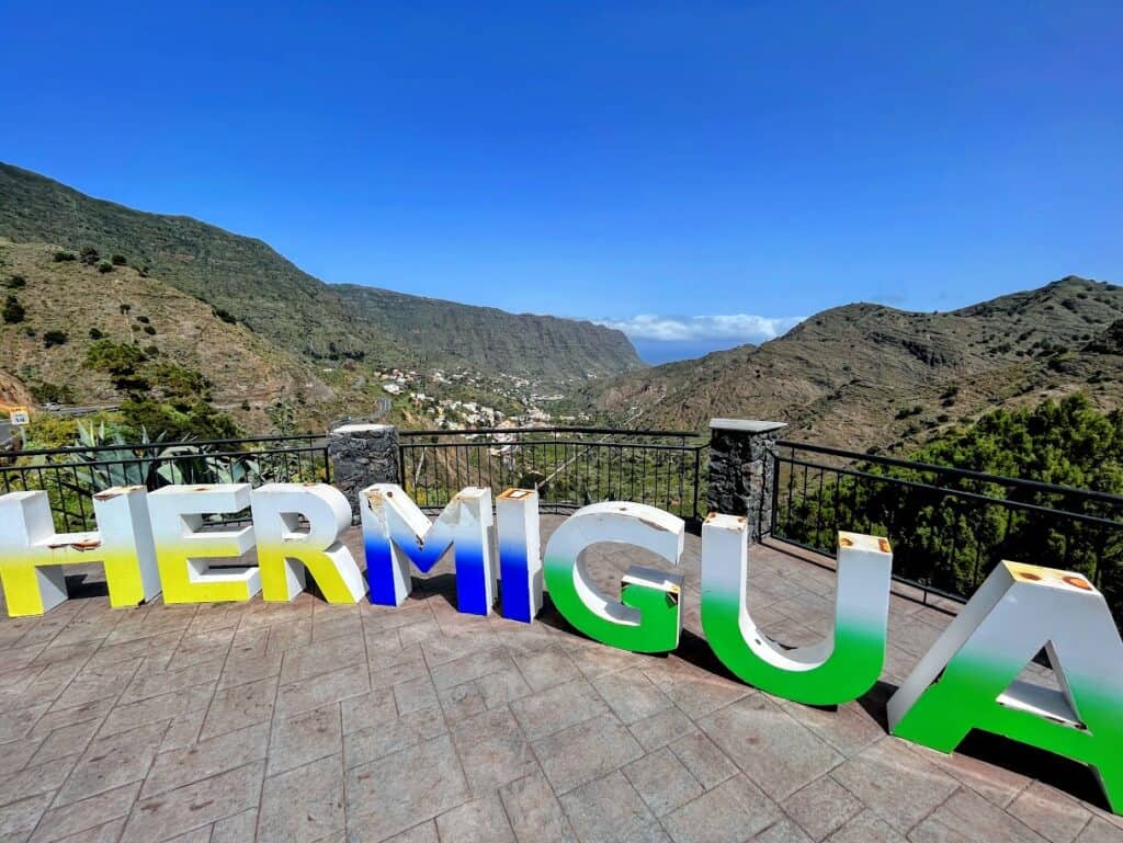 Brightly colored 'HERMIGUA' sign in bold letters stands out in a viewpoint overlooking the green valley and mountainous terrain of Hermigua.