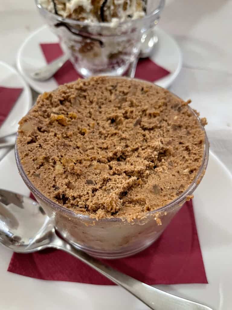 A dessert called Príncipe Alberto, a rich chocolate mousse topped with cookie crumbles, served in a small glass bowl with a spoon, on a table with a red napkin.