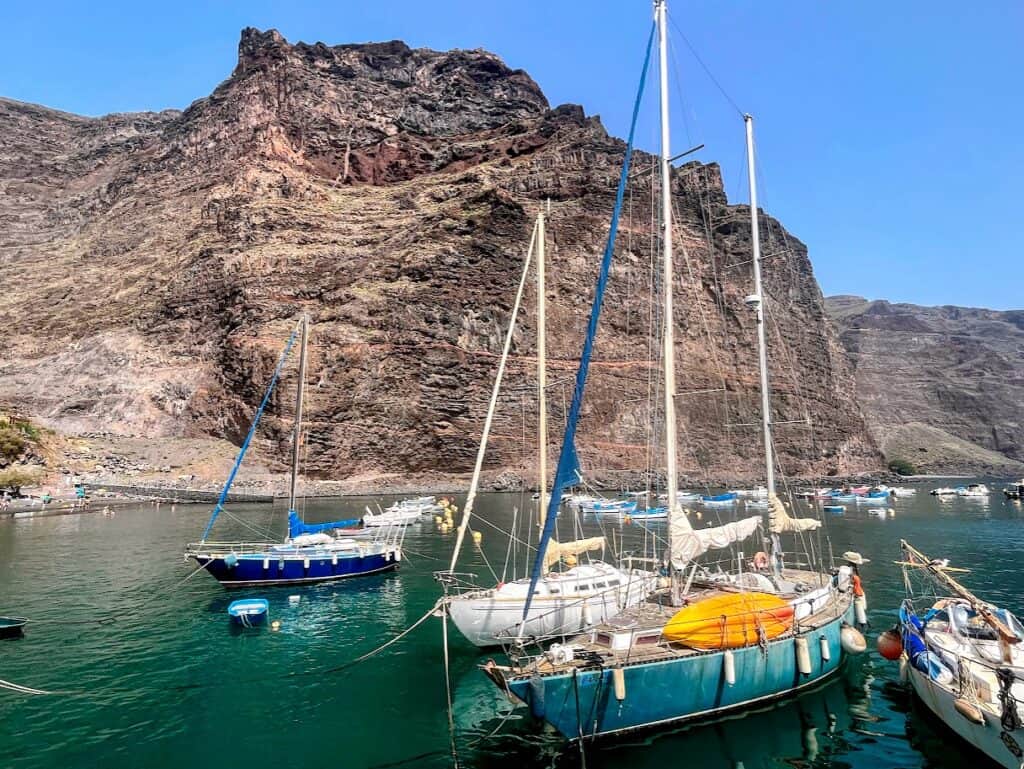 A tranquil marina with multiple sailboats moored in clear turquoise waters, set against a backdrop of rugged, towering cliffs under a clear blue sky.