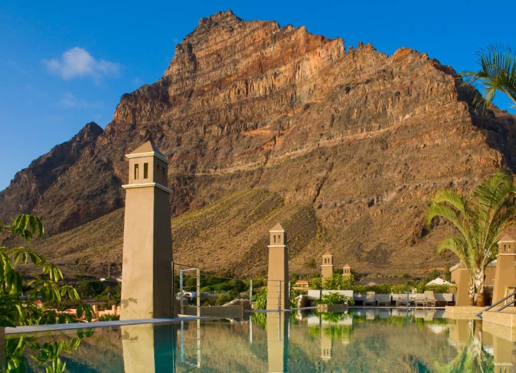 A serene pool with clear, reflective water at Hotel Playa Calera, set against the backdrop of a rugged mountain under a blue sky, lined with palm trees and a tower-like structure that complements the natural beauty of La Gomera, Canary Islands