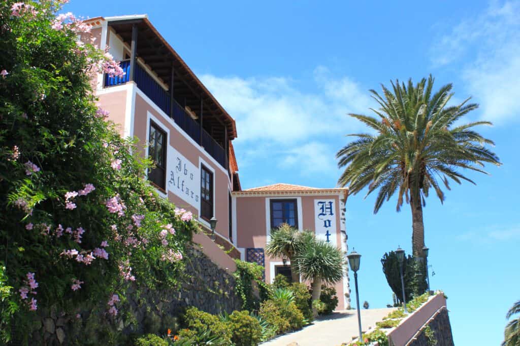 
This image shows Hotel Rural Ibo Alfaro in Hermigua, La Gomera, Canary Islands. The hotel is a charming, peach-colored building with white trim and a traditional Spanish architectural style. It is perched on a hillside and framed by lush greenery, pink blooming flowers, and a tall palm tree. A clear blue sky provides a bright background, highlighting the hotel's picturesque setting.