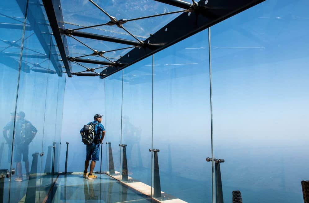 A lone traveler gazes out from the transparent walls of Mirador de Abrante in La Gomera, surrounded by reflections and the boundless blue of sea and sky.