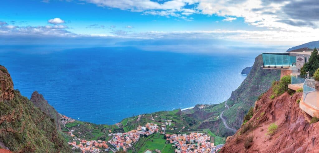 A panoramic view from Mirador de Abrante in La Gomera, showing the glass walkway jutting out over the cliff, offering visitors a dramatic perspective of the landscape.