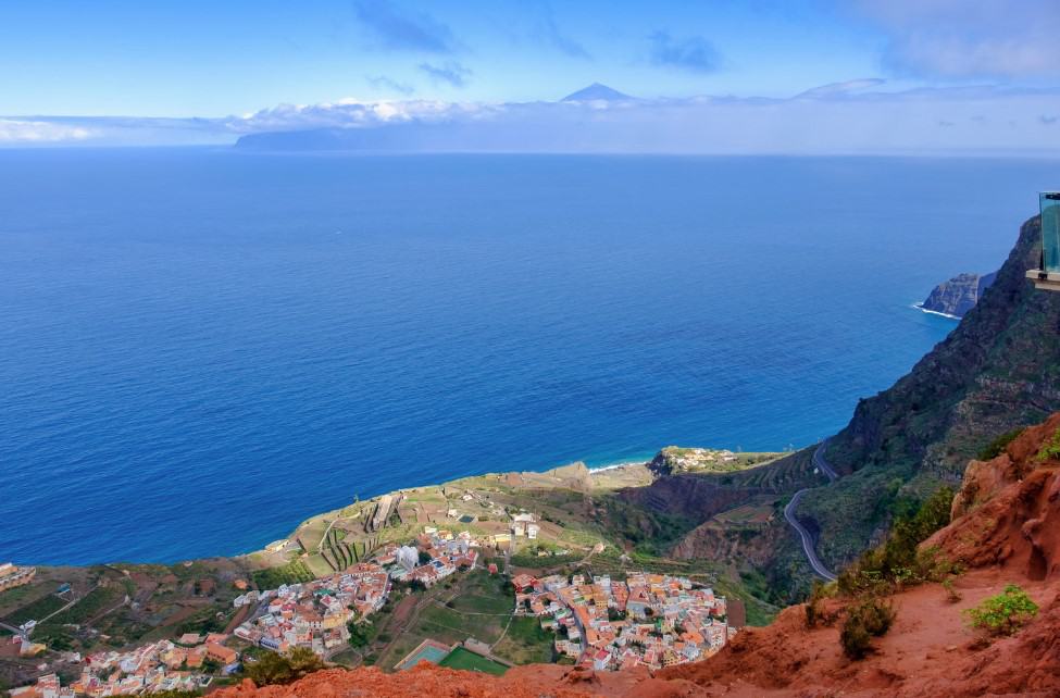 From Mirador de Abrante in La Gomera, the majestic Mount Teide of Tenerife looms in the distance, rising above the clouds and framed by the deep blue sea.