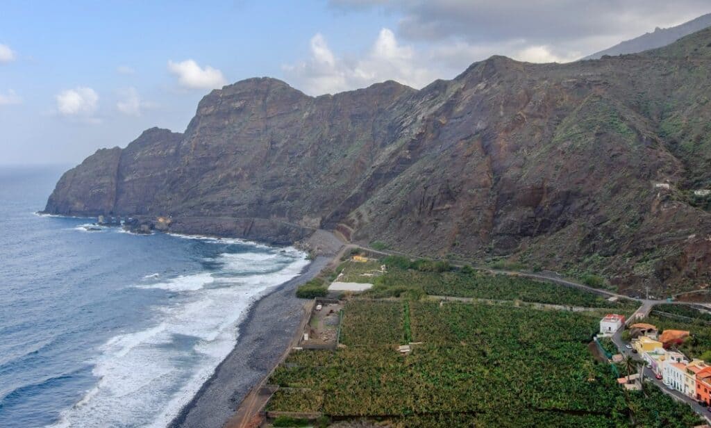 Aerial view of a rugged coastline with towering cliffs on one side and the pebble-strewn Hermigua beach bordering banana plantations on the other.