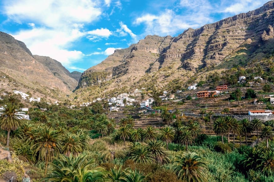 A panoramic view of La Gomera's lush Valle Gran Rey, with terraced hillsides, palm trees, and white houses nestled at the foot of tall, rugged cliffs under a blue sky.