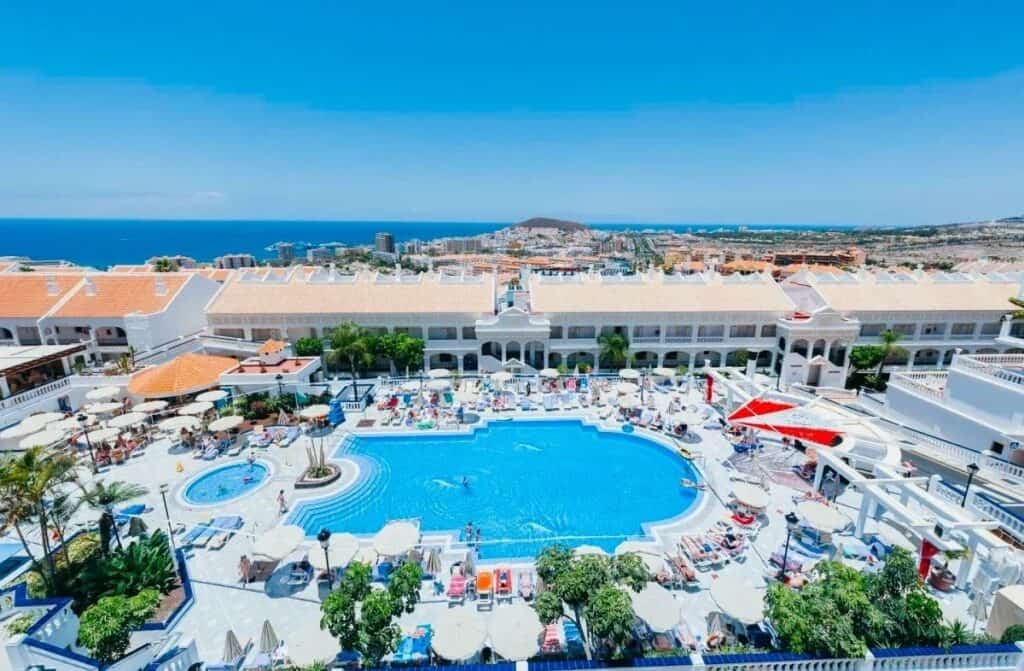 A bustling resort pool at Hollywood Mirage in Tenerife with guests lounging under umbrellas, flanked by white buildings with terracotta roofs, overlooking a coastal cityscape with the ocean horizon in the background.