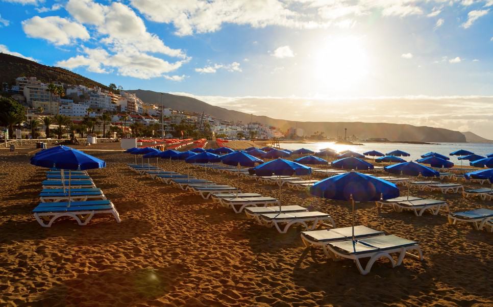 A stunning view of Playa de Los Cristianos in Tenerife, with rows of blue sun loungers and umbrellas on the sandy beach, perfectly capturing the essence of Los Cristianos hotels on the beach.