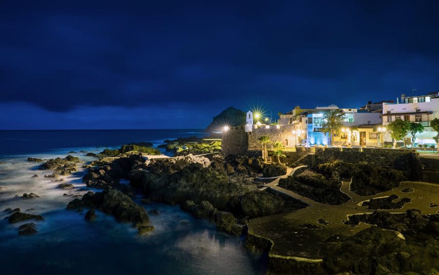 A stunning nighttime view of a coastal village in Tenerife, with twinkling lights reflecting off the water and rocky coastline, highlighting one of the many reasons to visit Tenerife.