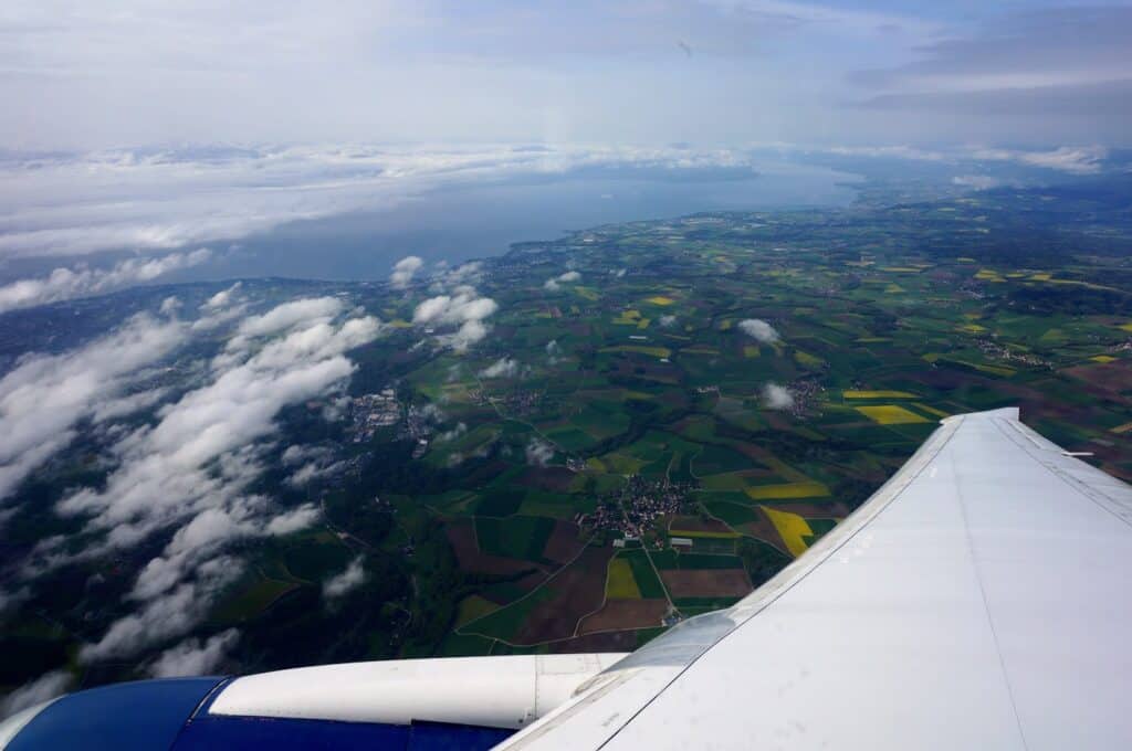 An aerial view from an airplane window as it descends over Geneva, Switzerland, revealing a patchwork of green fields, small towns, and the expansive Lake Geneva surrounded by clouds. This marks the beginning of an exciting journey through the city’s must-see sights in a 2-day itinerary.