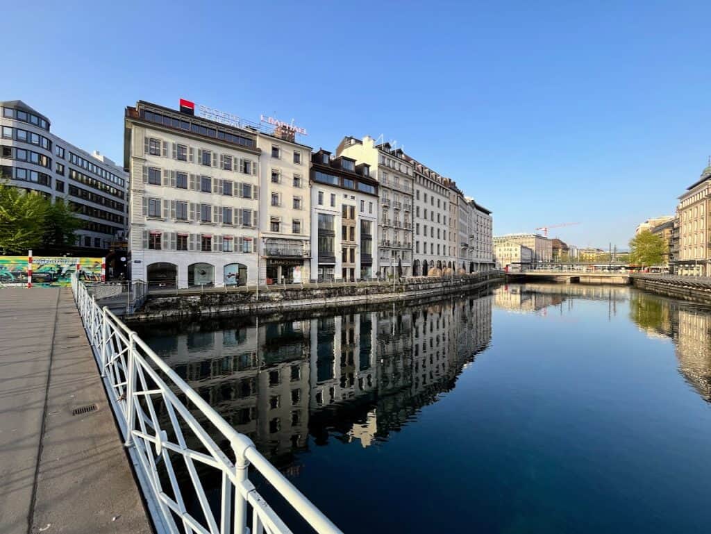 A tranquil view of Geneva's waterfront with the calm waters of the Rhône River reflecting the city's architecture, a serene spot to visit during your one day in Geneva.
