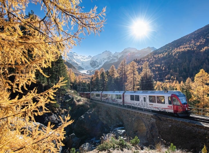 A scenic train crossing a bridge in the Swiss Alps during fall, surrounded by golden trees and snow-capped mountains in the distance. One of the best Lake Geneva, Switzerland Things to Do is experiencing the region’s beauty by scenic railway.