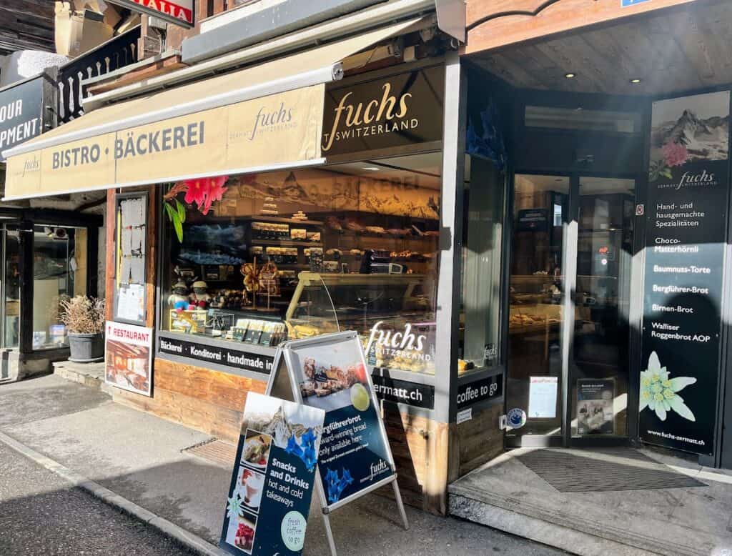 The inviting facade of Bäckerei Fuchs showcases its selection of baked goods, with tempting pastries and bread displayed in the window. The charming Swiss bakery has a rustic design that draws in locals and visitors alike. Stopping here for a treat is a delightful addition to any Zermatt adventure.