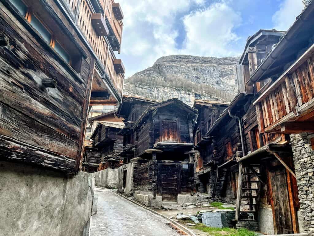 A narrow cobblestone path winds through the historic wooden buildings of Hinterdorfstrasse in Zermatt, showcasing traditional Swiss architecture against a backdrop of towering mountains. The rustic structures, some with ladders leading to upper levels, reflect the charming character and rich history of this picturesque village.