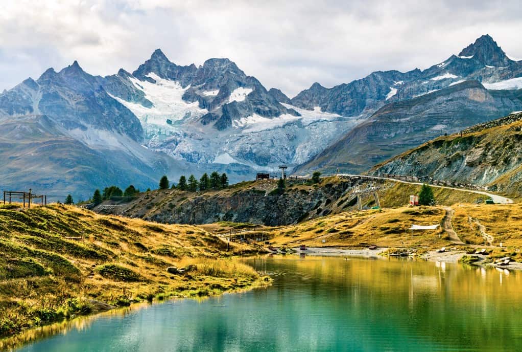 A tranquil view of Lake Leisee surrounded by lush greenery, with the iconic Matterhorn towering majestically in the background. The lake's clear waters reflect the mountain and the vibrant blue sky.