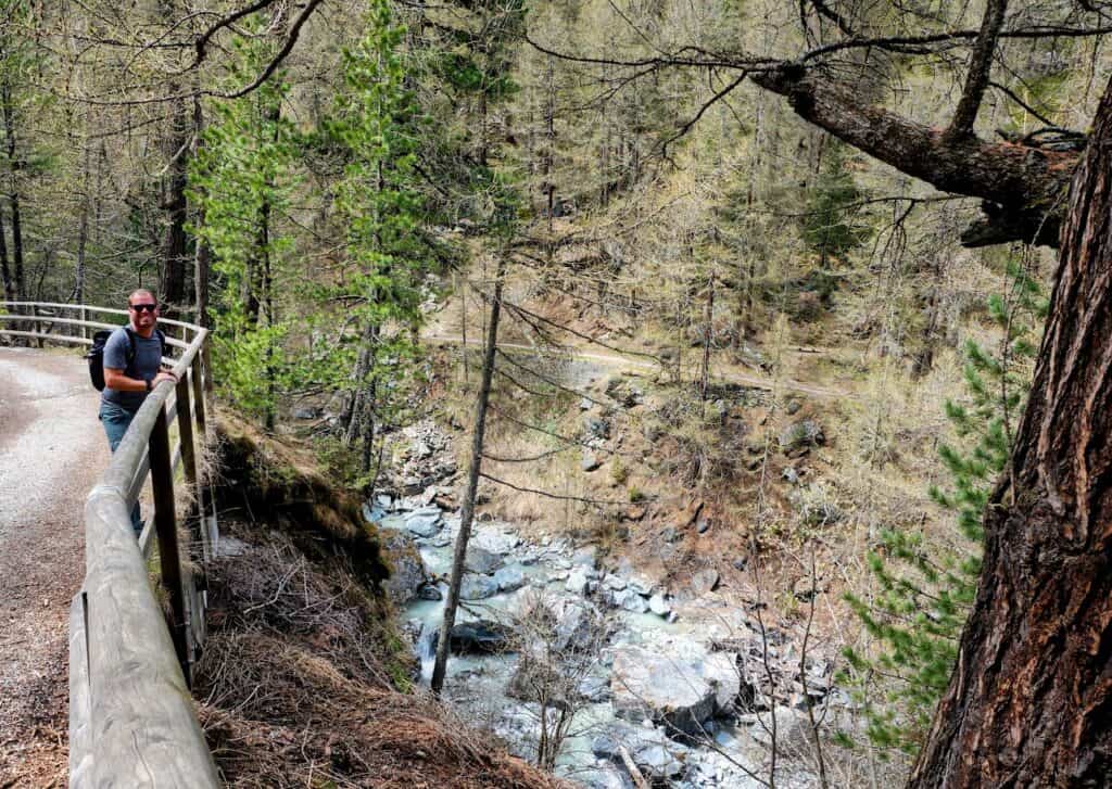 Nicholas stands along a scenic trail by the river in Zermatt, surrounded by towering trees and rocky outcrops. The clear blue water of the river flows gently, reflecting the natural beauty of the area as he enjoys a peaceful moment in nature.