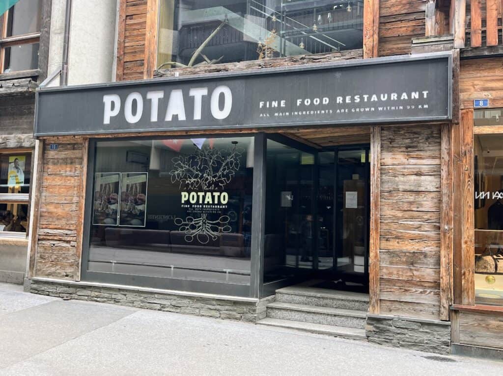 The exterior of Potato Fine Food Restaurant in Zermatt features a sleek, modern design with large glass windows showcasing a stylish interior. The restaurant's sign is prominently displayed, and the wooden facade adds a rustic touch. Enjoying a meal here is a highlight of any Zermatt itinerary.