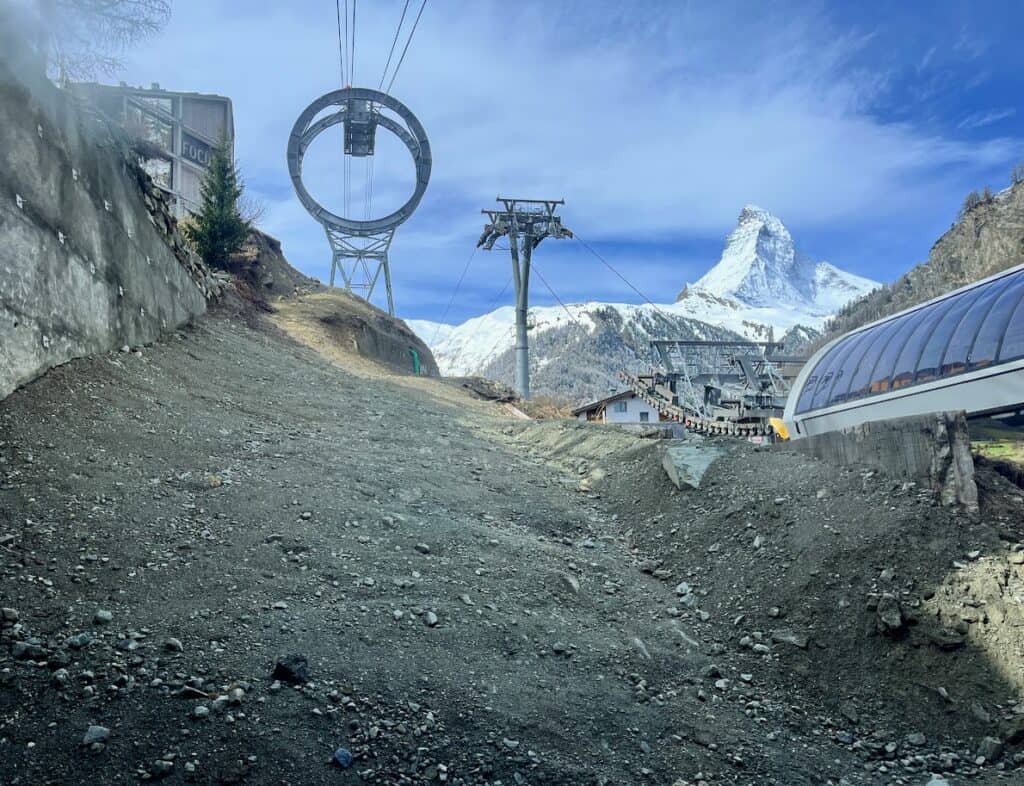 A captivating view from the base of a gondola lift in Zermatt, showcasing the iconic Matterhorn in the background. The image features the gondola's sleek design ascending the mountain, surrounded by rocky terrain and patches of grass, while the clear blue sky provides a stunning contrast to the snowy peaks above.