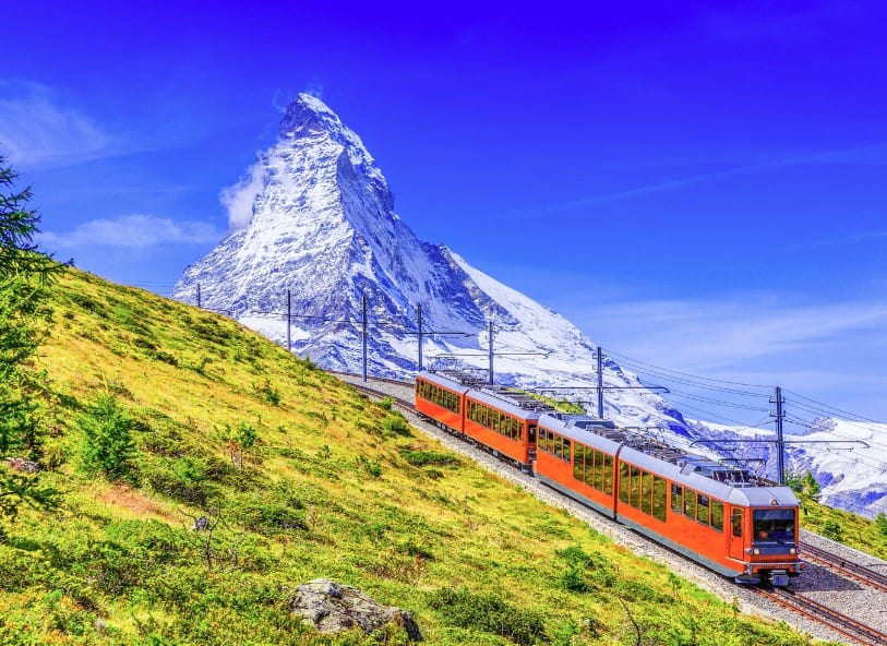 A bright orange train climbs a green mountainside with the Matterhorn towering in the background. Scenic rides like this show exactly why the answer to is Zermatt worth visiting in summer is yes.