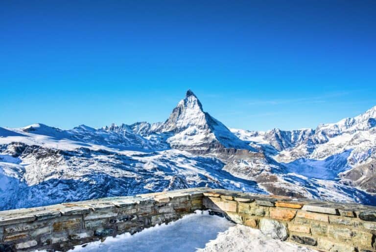 The viewpoint at Gornergrat displays a panoramic view of the Matterhorn and surrounding peaks. Tourists admire the breathtaking scenery, with the vibrant blue sky contrasting beautifully against the snow-capped mountains, showcasing the magic of Zermatt.