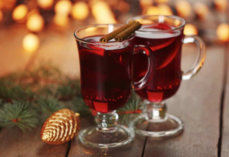 Two glasses of warm mulled wine with cinnamon sticks, set against a festive backdrop of pine branches, glowing lights, and a cozy atmosphere perfect for the winter season.